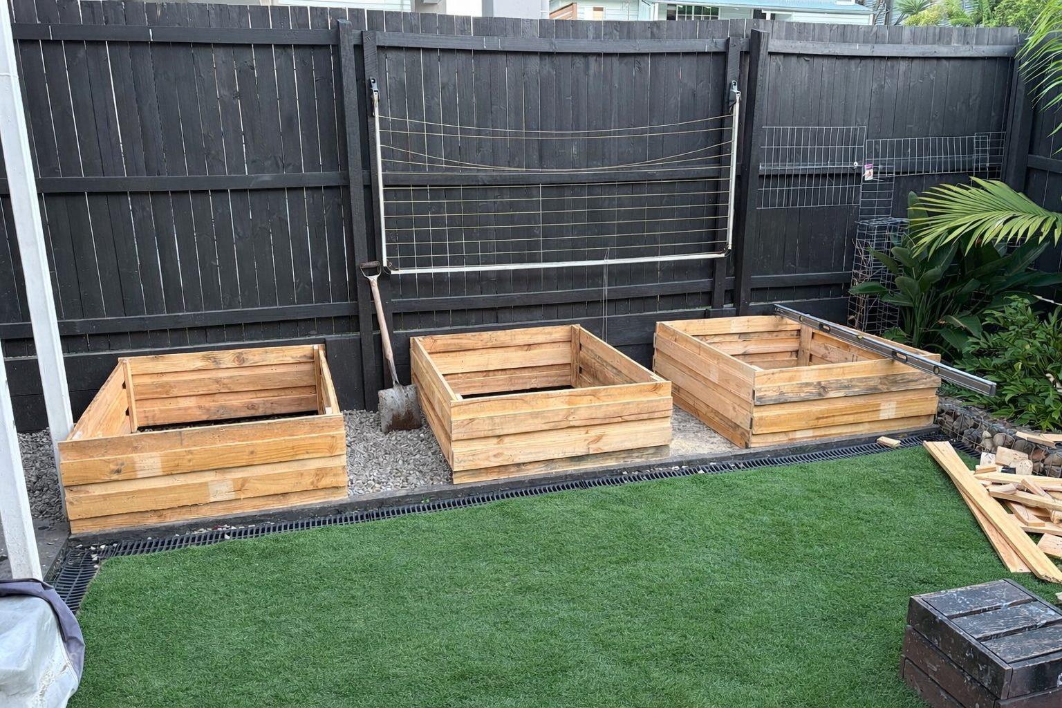 Three empty treated-timber raised garden beds built and set out on a gravel strip along the dark-stained back fence, lawn in front, timber offcuts still on the grass