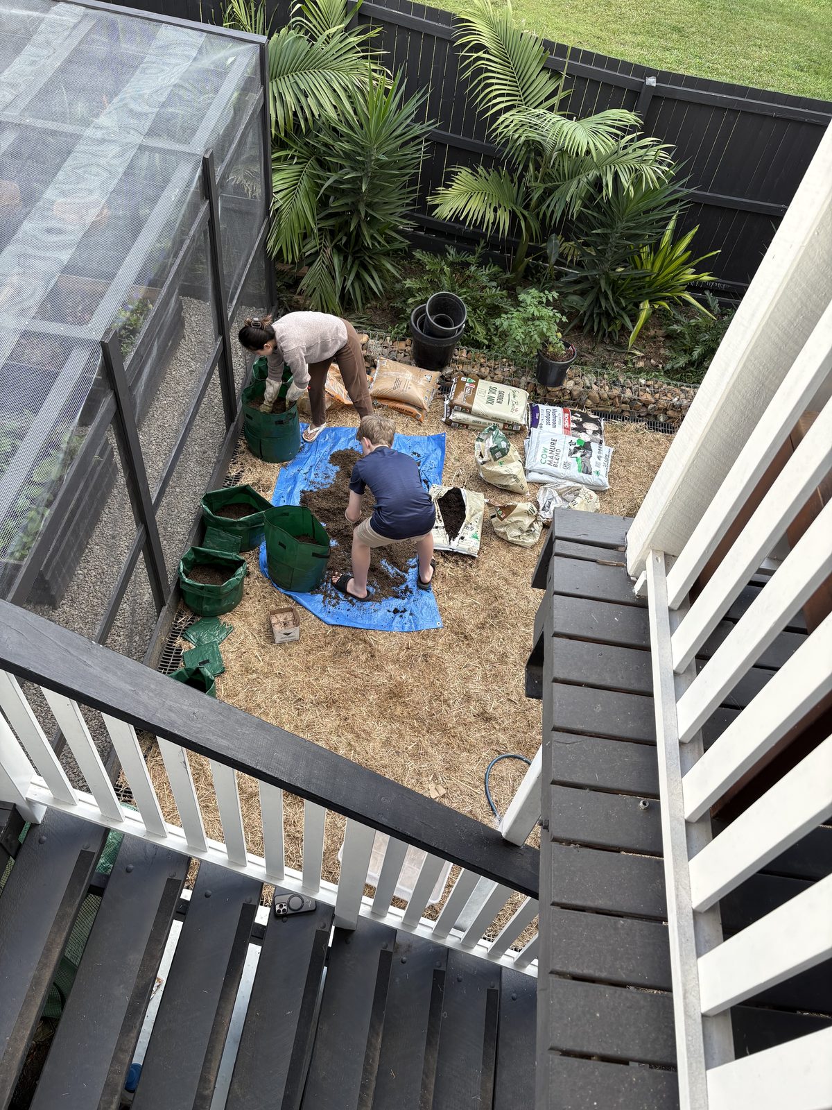 Overhead view of two people mixing soil, compost and manure on a blue tarp and filling green fabric grow bags, with bags of garden soil mix, mushroom compost and cow manure blend lined up alongside